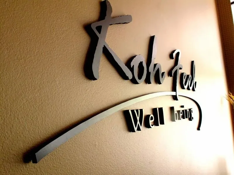 3D metal Koh Feel Well Being script wordmark on a warm tan wall
